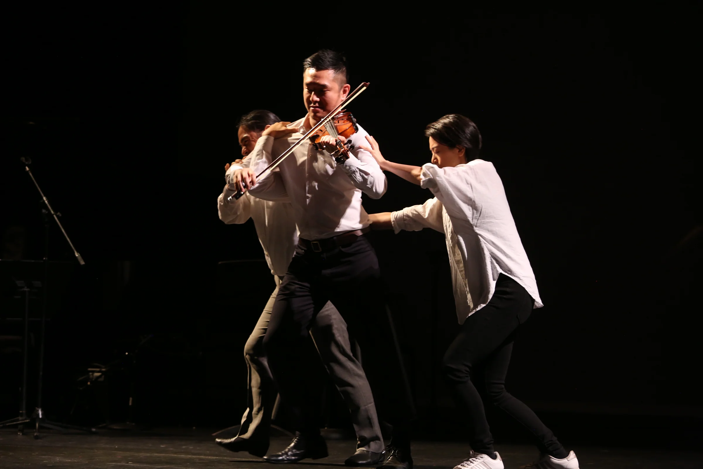 A violinist performing as movement artists reach toward him on stage